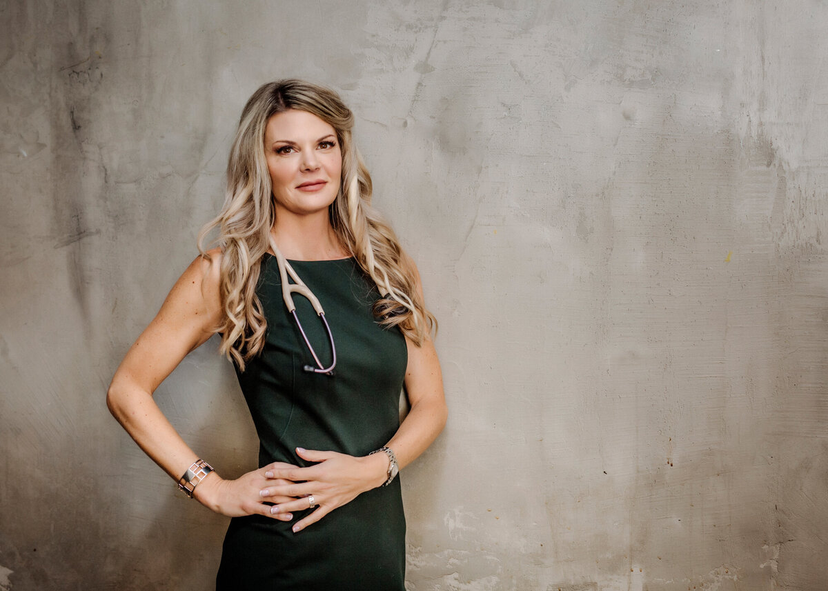 A female nurse practitioner wearing a green dress & stethoscope leaning against a grungy beige wall while posing for a South Shore MA branding photographer