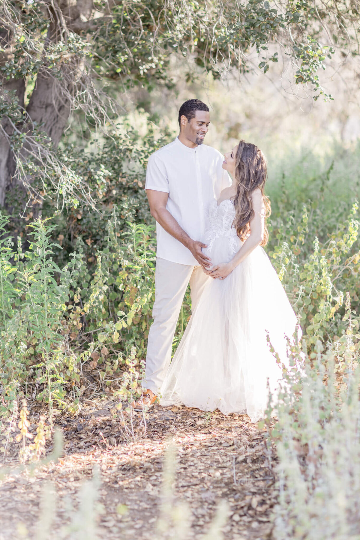A couple stands lovingly in a sunlit garden. The woman, wearing a flowing white dress, holds her pregnant belly as the man, in a white shirt and light pants, embraces her under the dappled shade of trees.