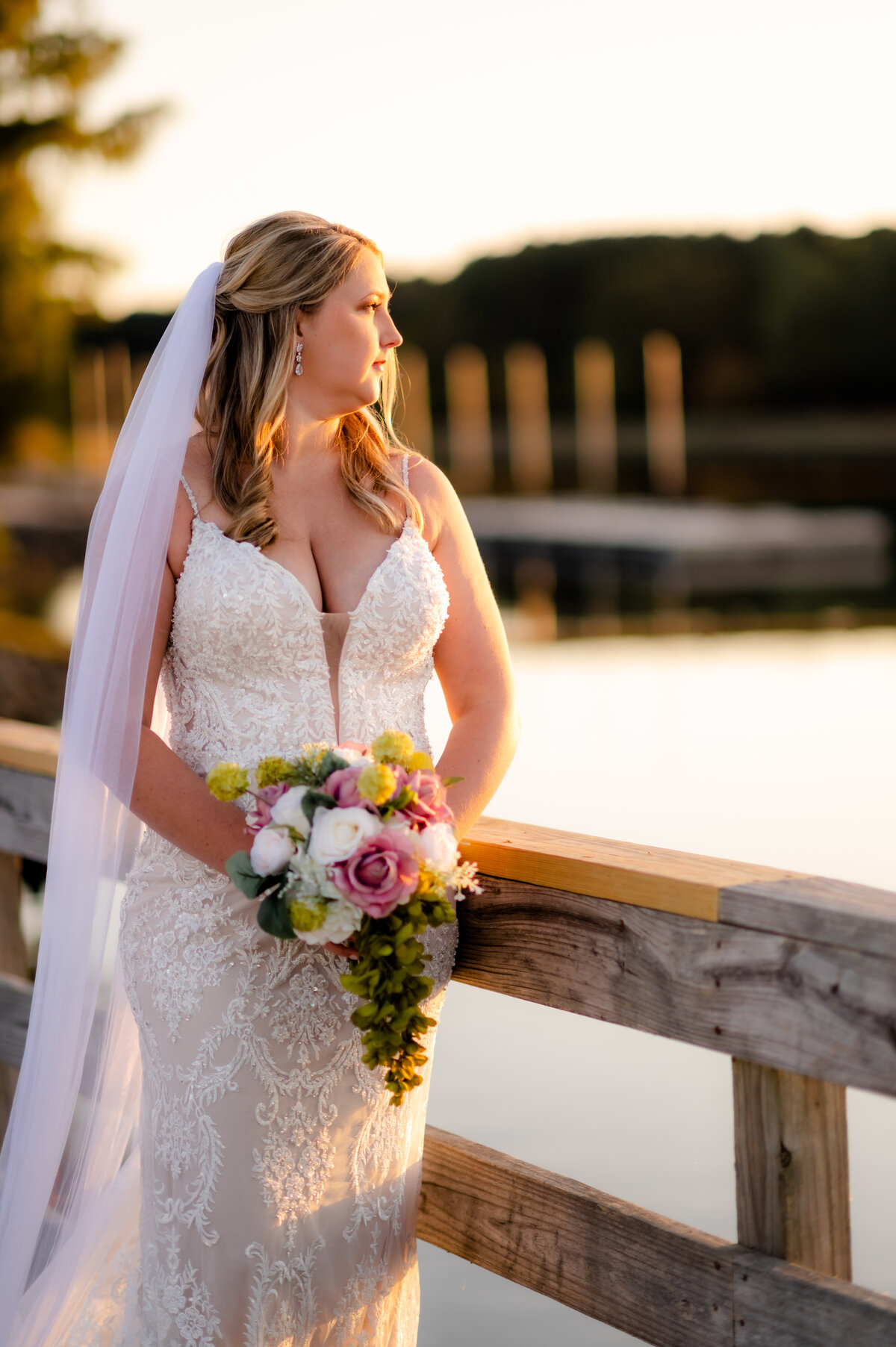 sun setting over a bride as she looks over the lake's water as she holds a small pink rose bouquet