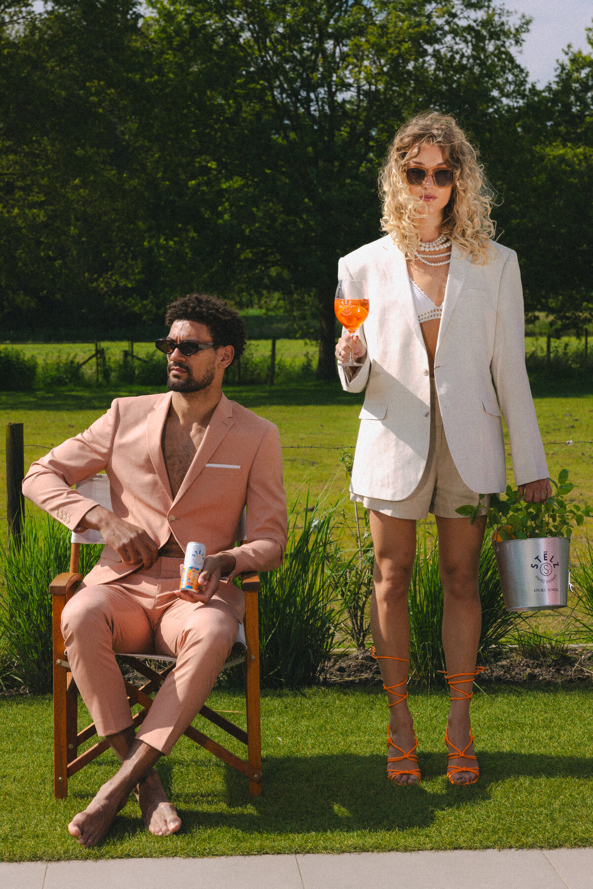 A stylish couple dressed in high fashion, seated outdoors with a STËLZ Classic Spritz in hand. The man wears a peach-colored suit, while the woman stands confidently, holding a glass of spritz, exuding elegance and luxury. Photography by Bo Eppink.A stylish couple dressed in high fashion, seated outdoors with a STËLZ Classic Spritz in hand. The man wears a peach-colored suit, while the woman stands confidently, holding a glass of spritz, exuding elegance and luxury. Photography by Bo Eppink.
