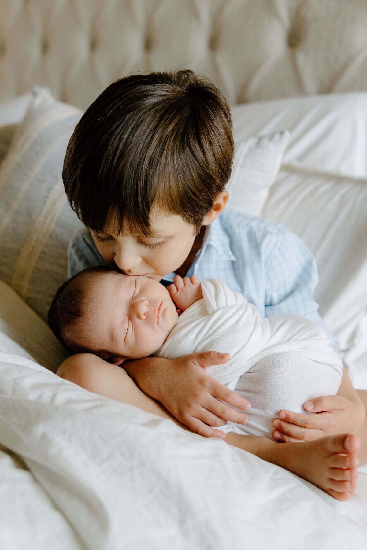 toddler boy holding baby brother while giving newborn a sweet kiss on cheek