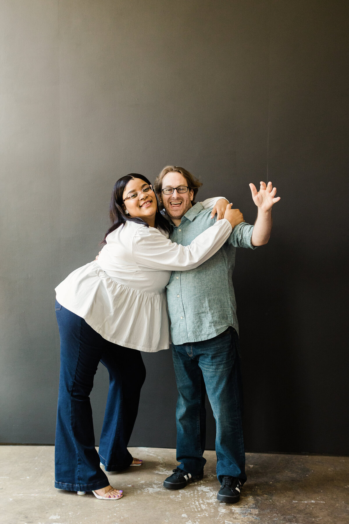 A couple playfully posing in front of a black background during their studio engagement photoshoot in Dallas, Texas. The woman on the left is wrapping her arms around the man on the right. She is wearing a long sleeve, white blouse, jeans, and glasses. The man is wearing a short sleeve dress shirt, jeans, and glasses.