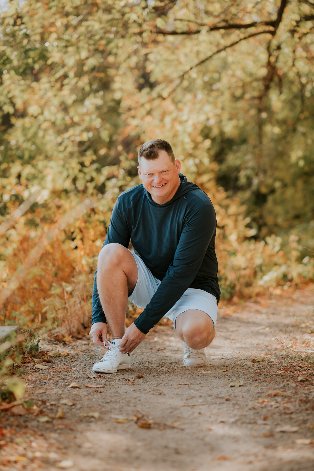 Natural light dating profile photo of a man in activewear tying his shoes on a wooded trail in Minneapolis, captured for an active online dating profile.