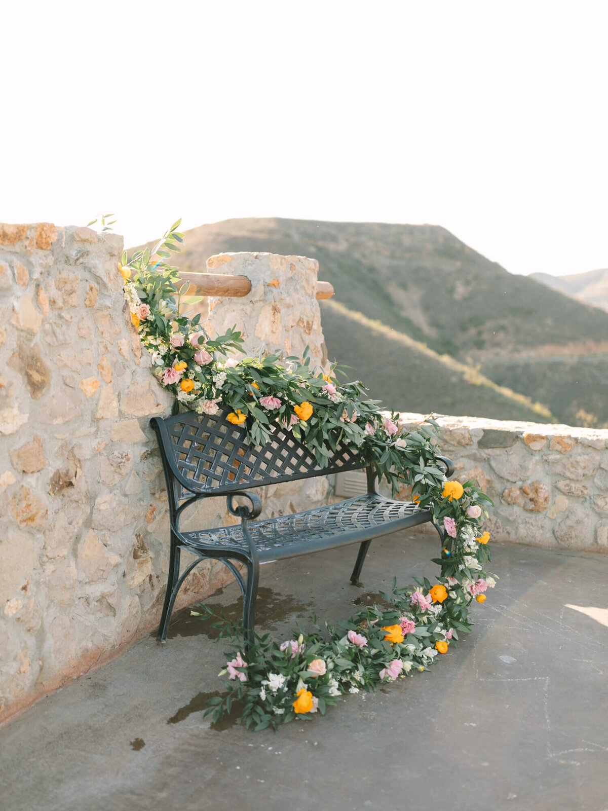 Ornate black bench adorned with a garland of vibrant flowers against a rustic stone wall.