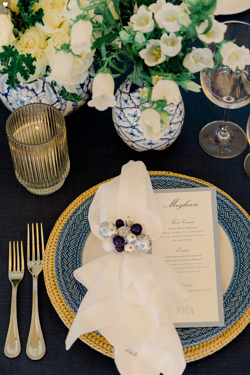 Menu with blue border sitting on blue knot plate and gold charger. Forks to the left and table design with blue and white vases and white roses