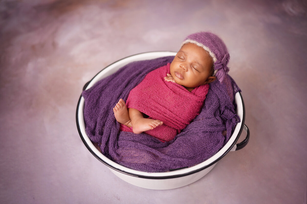 Black newborn baby girl with a purple knit hat and magenta swaddle.  The babies hands and feet are exposed.  She is laying on a purple blanket in a vintage white metal tub.  She is on a pink background.