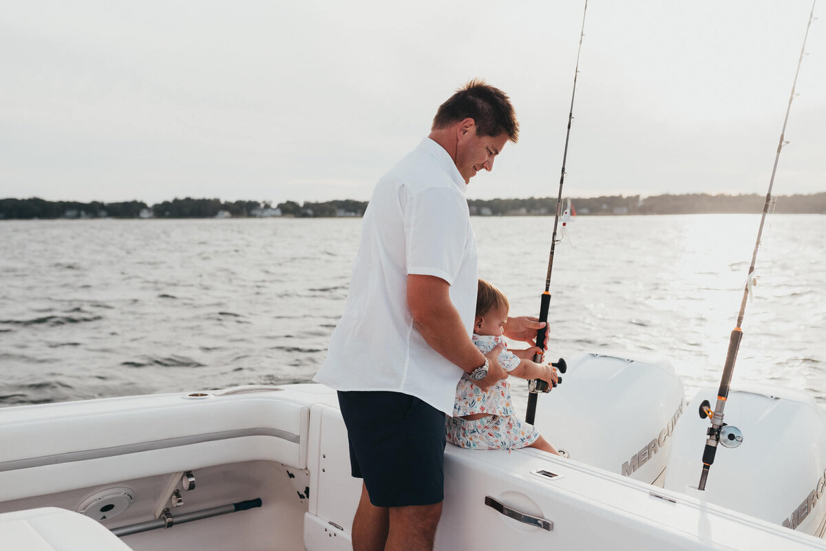 A father holds on to his young son who is holding one of his deep sea fishing rods on their boat at sunset