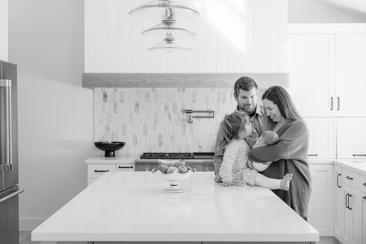 Candid black and white image of a family gathered in their kitchen during an in-home lifestyle photography session, captured by Suzanne Karp from Little Palm Photography in Northern California.