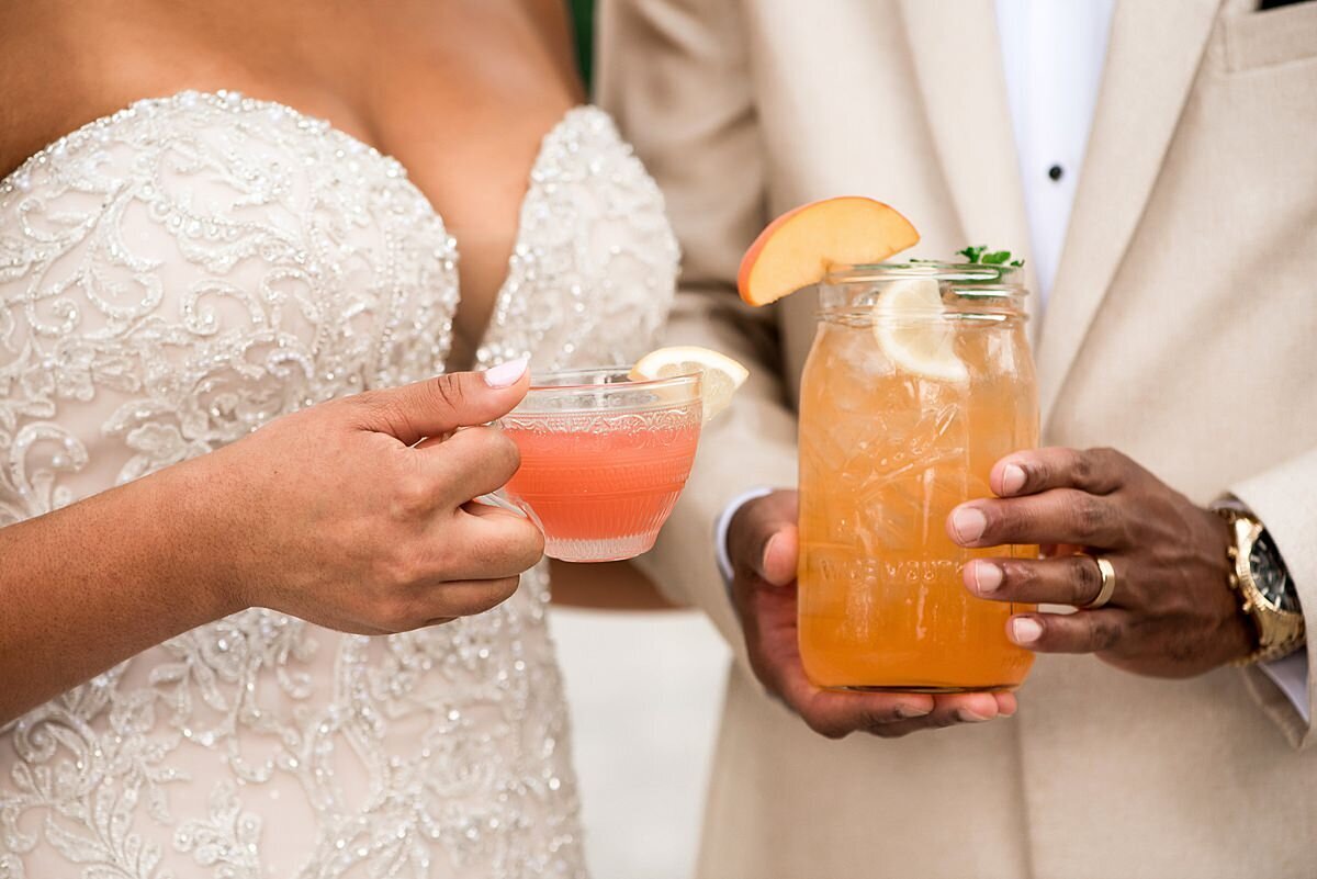 The bride, wearing a lace wedding dress with a sweetheart neckline holds a cut crystal punch glass of southern wedding punch. The groom, wearing a white tuxedo jacket, holds a signature summer peach tea cocktail at Ravenswood Mansion