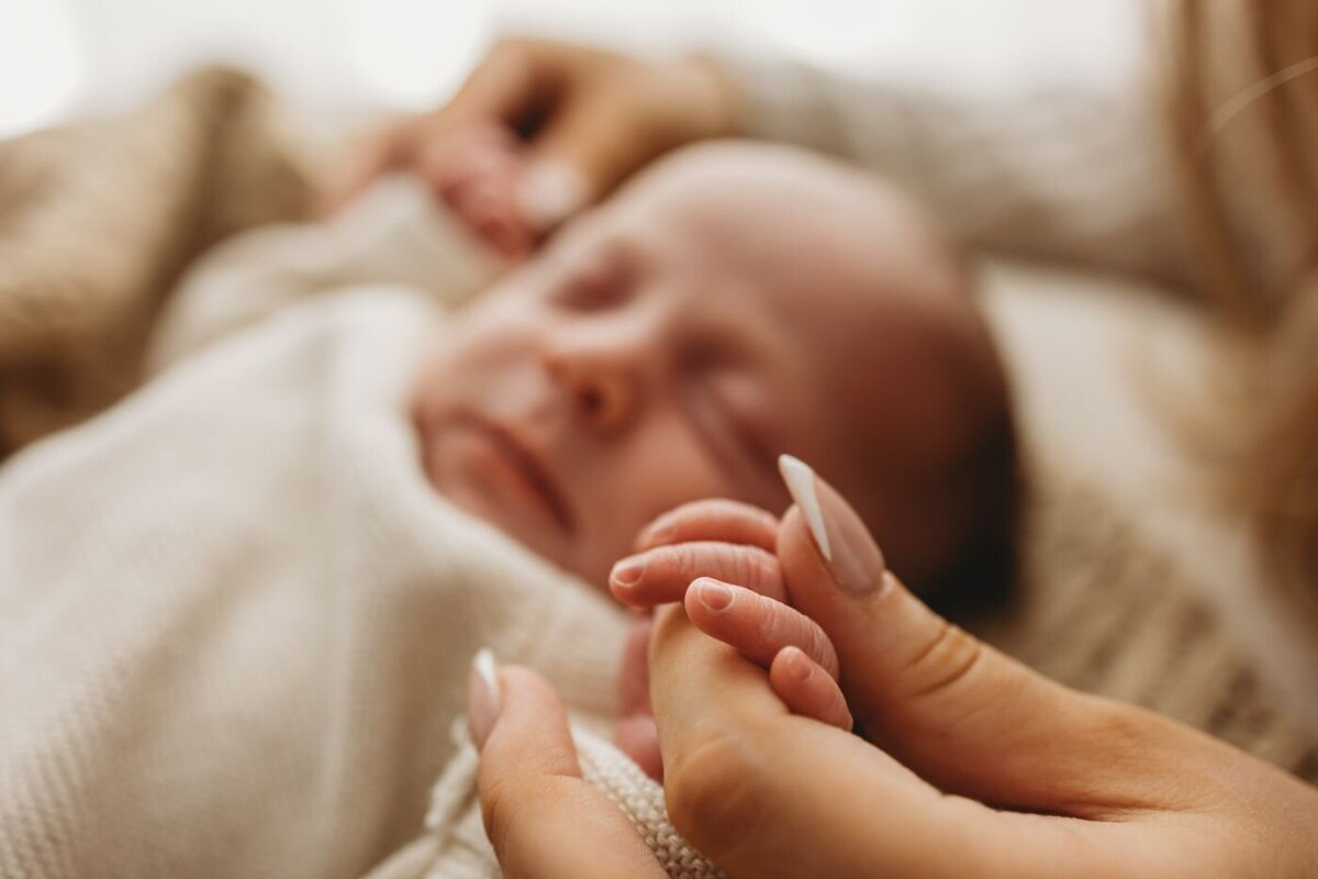 Intimate macro shot of a mother's manicured hand delicately holding the small hand of her sleeping newborn baby, who is wrapped in a textured cream fabric. Ruaa Hamdan Photography