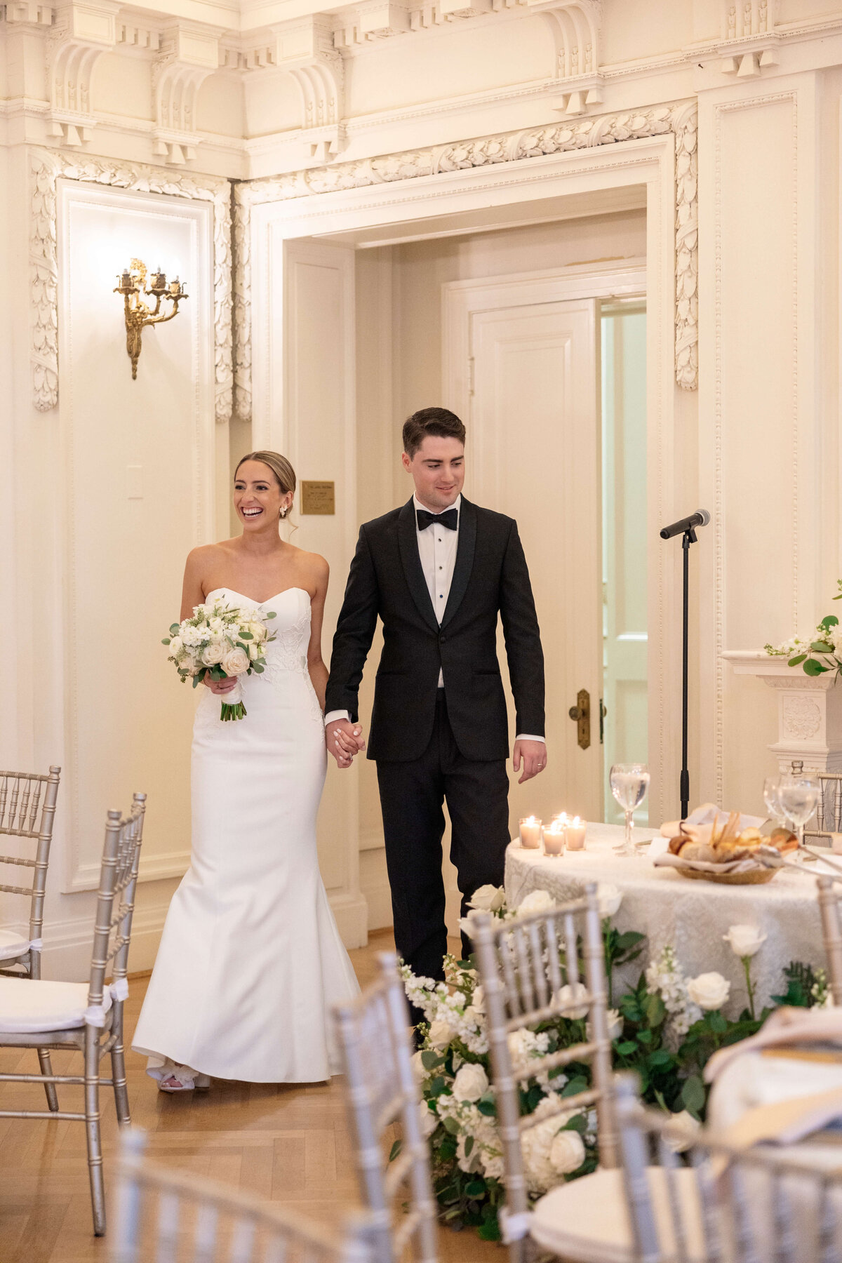 A bride in a white dress and a groom in a black tuxedo enter a decorated room hand in hand. The room features elegant white walls, floral arrangements, and set dining tables with candles. The bride holds a bouquet of white flowers.