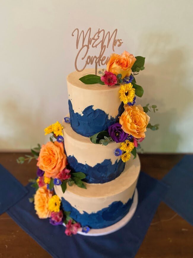 A tall tiered cake has tan frosting with navy waves and texture around the bottom of each tier. There's two large clusters of bright rainbow fresh flowers in two cascades down the sides.