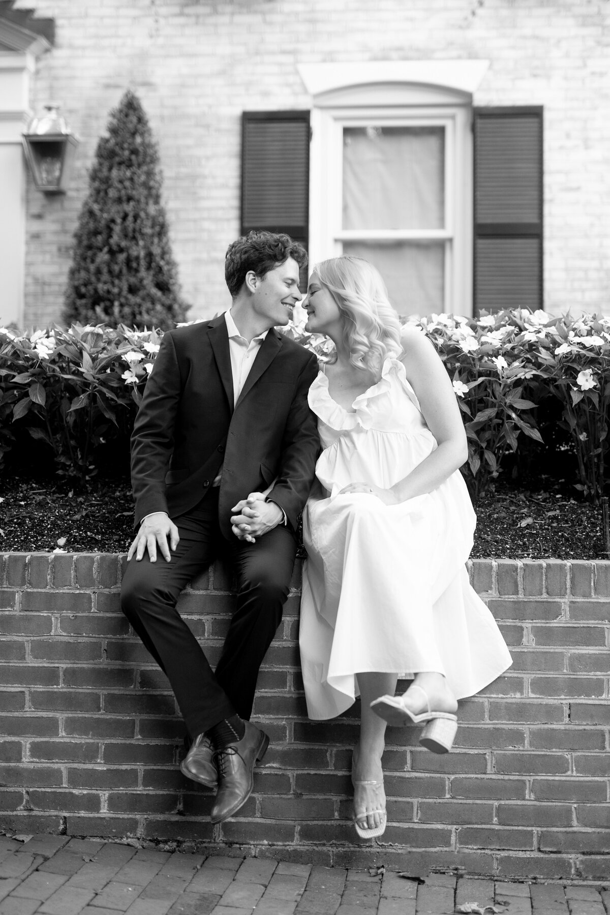 A couple sits on a brick wall, holding hands and smiling at each other. The woman is wearing a white dress, and the man is in a dark suit. Behind them are blooming flowers and a brick building with a window and shutters.