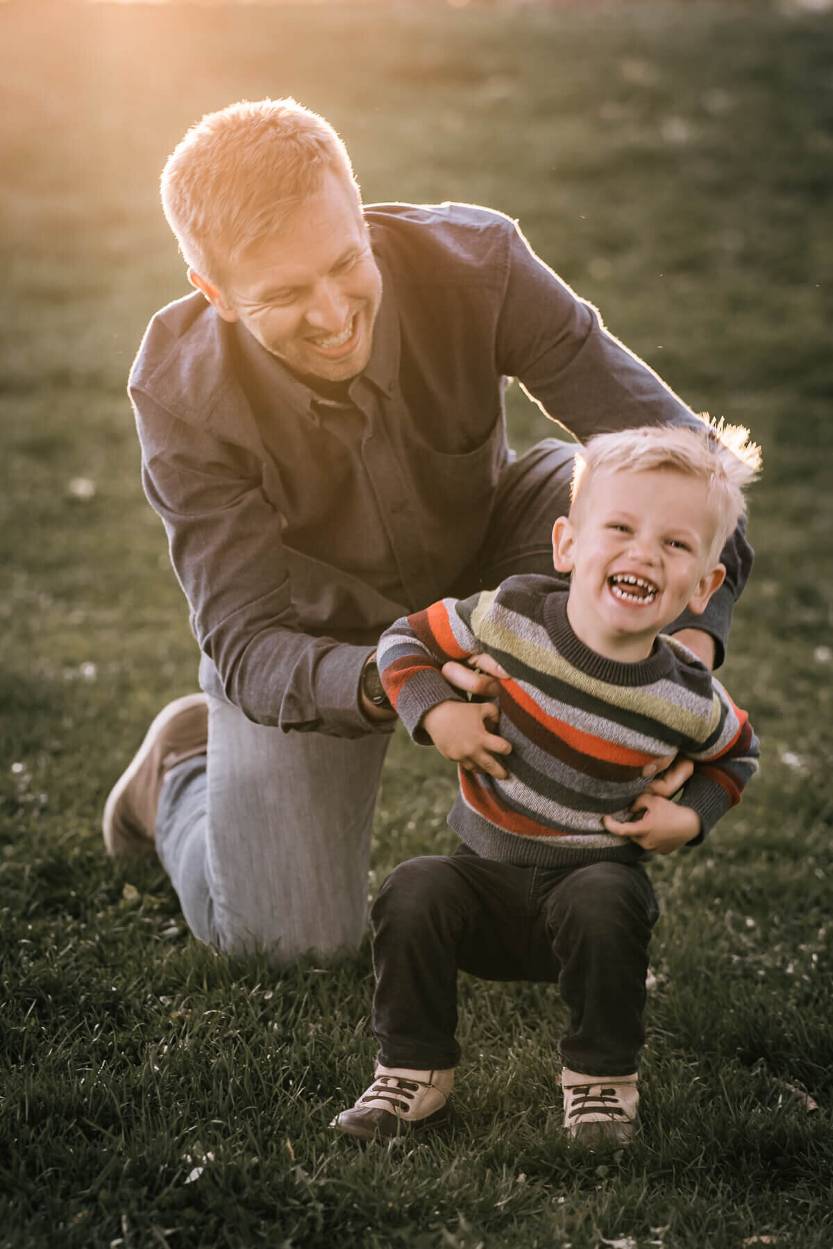 A small boy laughs as his father kneels down and tickles him from behind, as a sun flare illuminates the top left of the image.