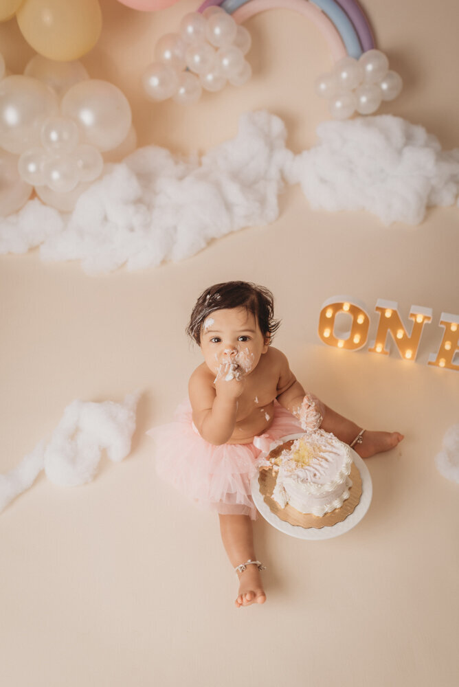 Photo shoot at Atlanta, Georgia cake smash photographer studio with one year old little girl in pink tutu smashing a cake with rainbow pastel balloon garland behind her with white fluffy clouds.