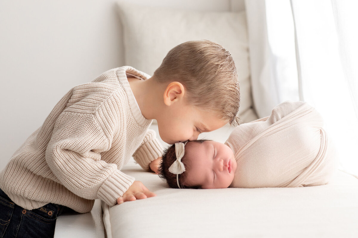 an older sibling kisses his new baby sisters head while she sleeps swaddled