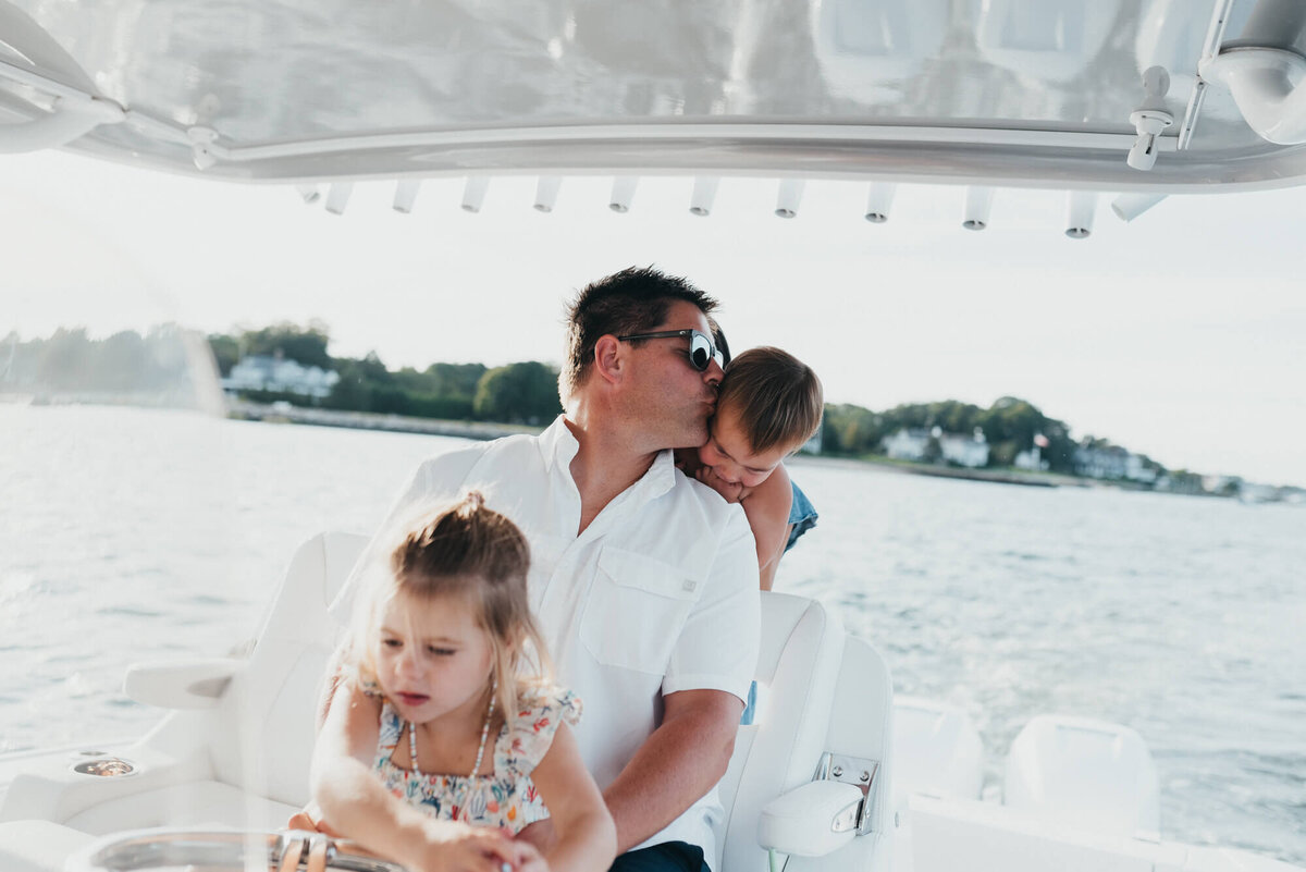 A father lets his daughter help him drive their boat while he gives his son a kiss over his shoulder