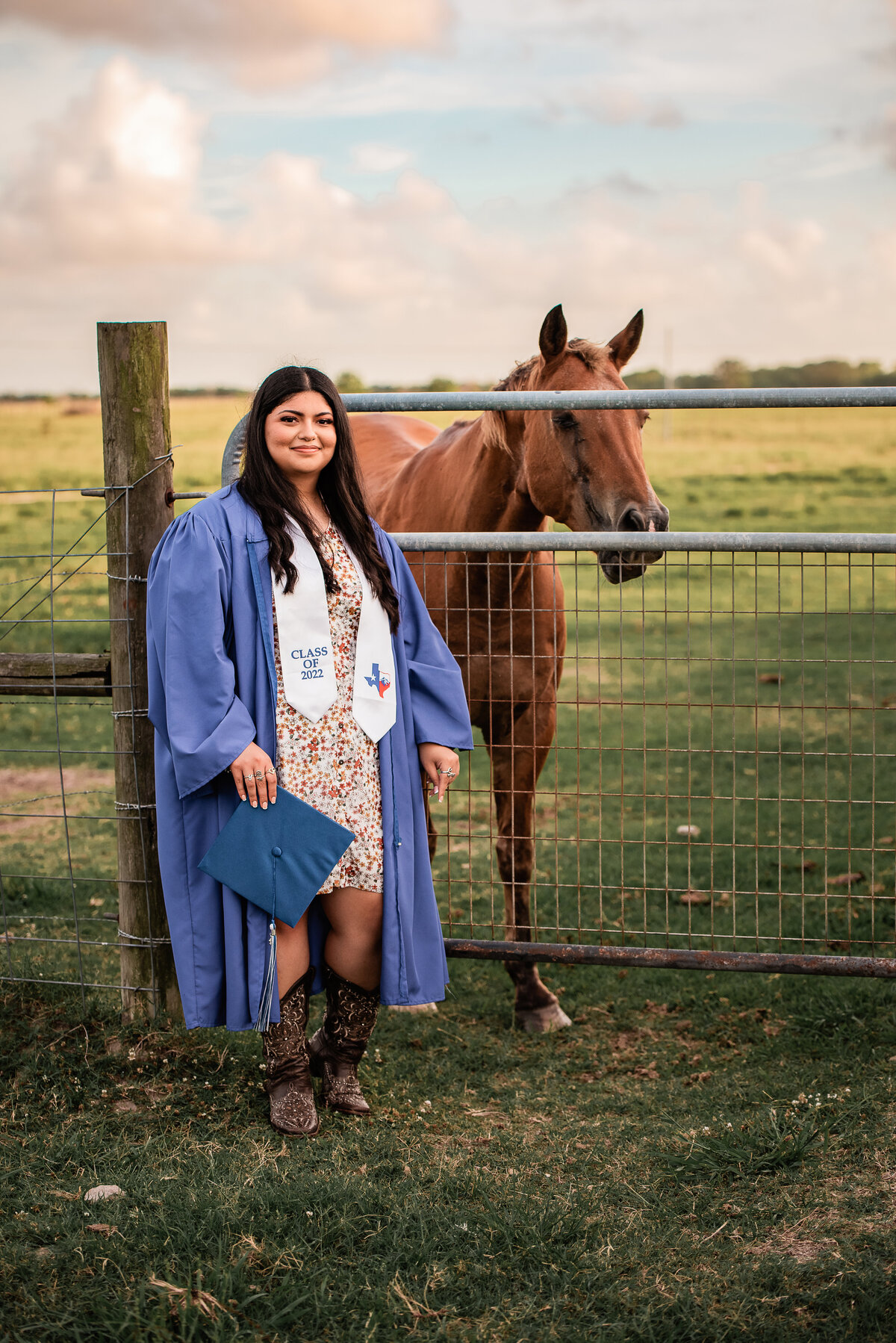 A girl wearing a blue cap and gown stands near a fence next to a horse.