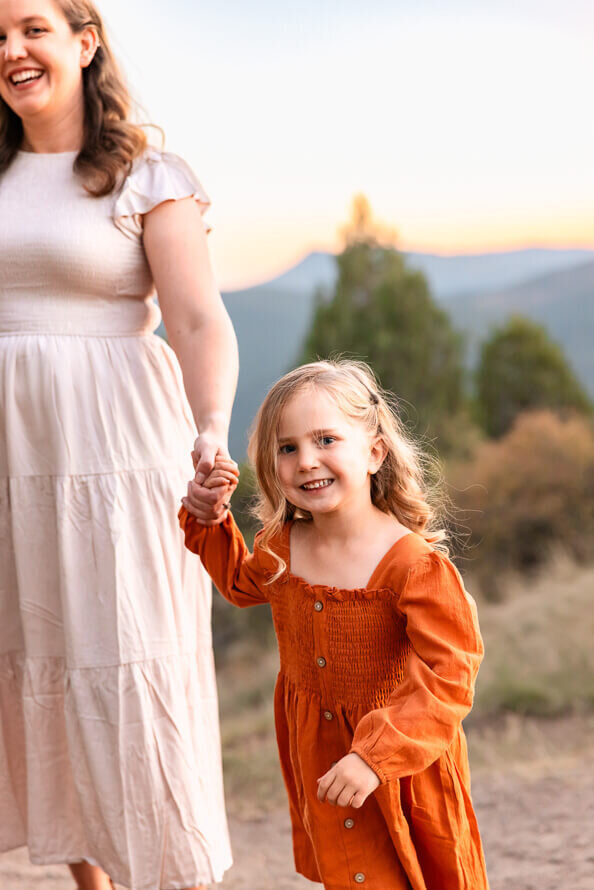 a little girl dressed in orange holds her mothers hand and smiles at camera.