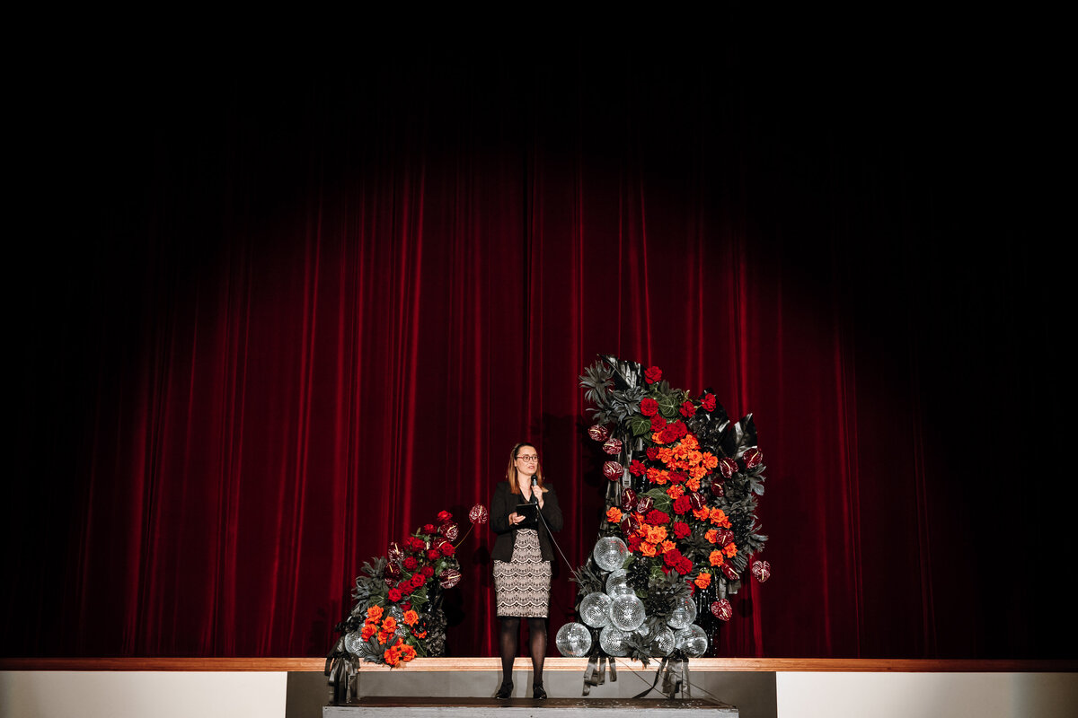 The MC standing in between two large floral arrangements on stage of regal cinema wedding.
