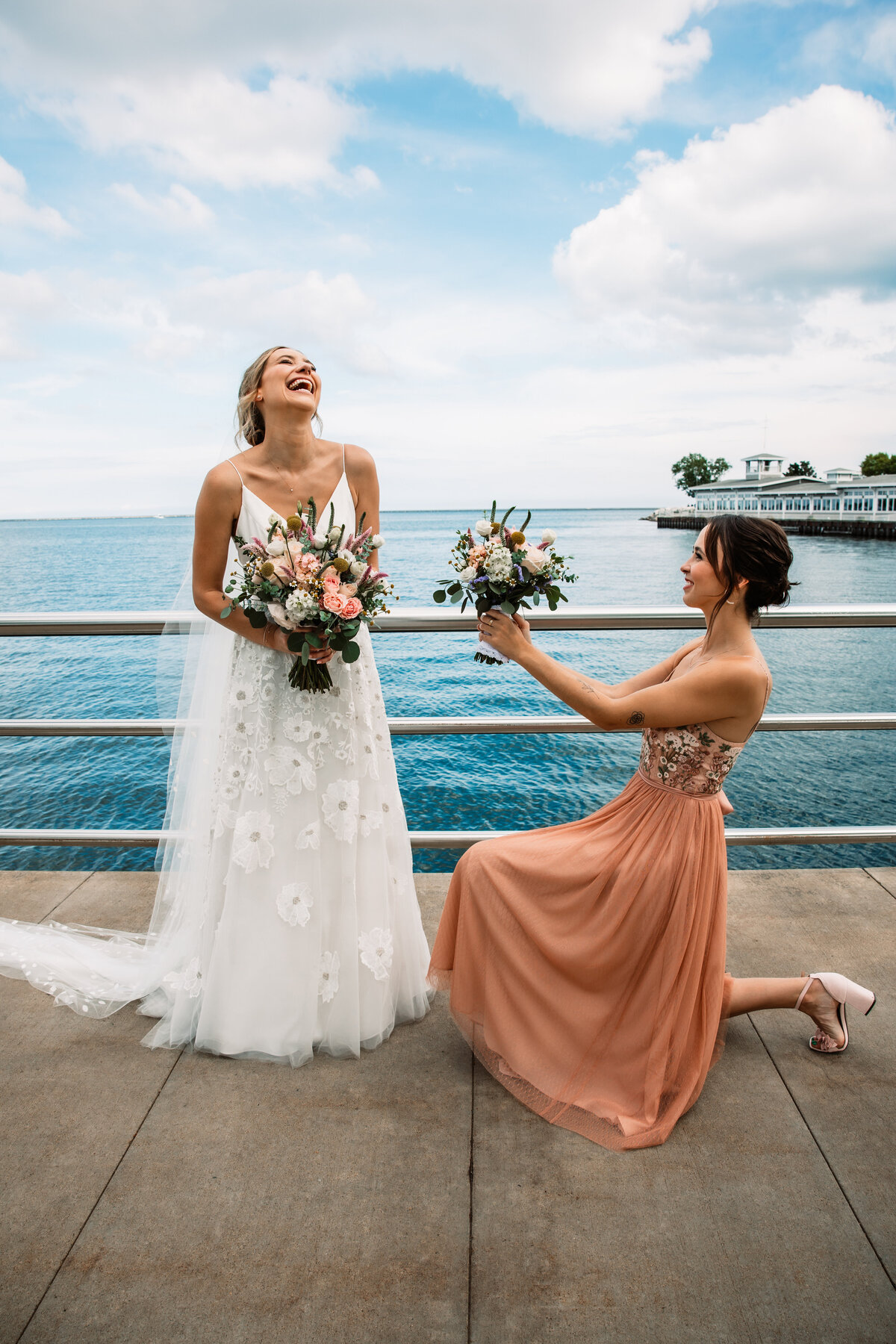 A bridesmaid in a pink dress gets on one knee on a dock and proposes to the bride with bouquet