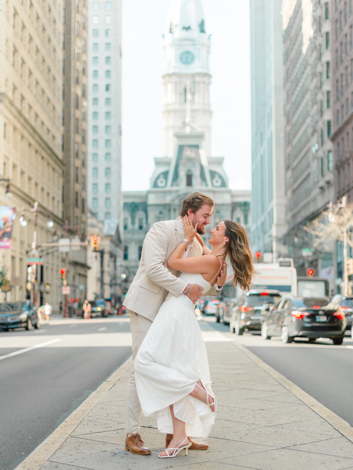 A couple shares a joyful, romantic moment in the heart of Philadelphia, with City Hall rising majestically behind them on Broad Street. The bride-to-be, in a flowing white dress and heels, is dipped back by her fiancé in a playful embrace. Captured in golden morning light by Elizabeth Kane Photography, a Luxury Philadelphia Wedding Photographer.