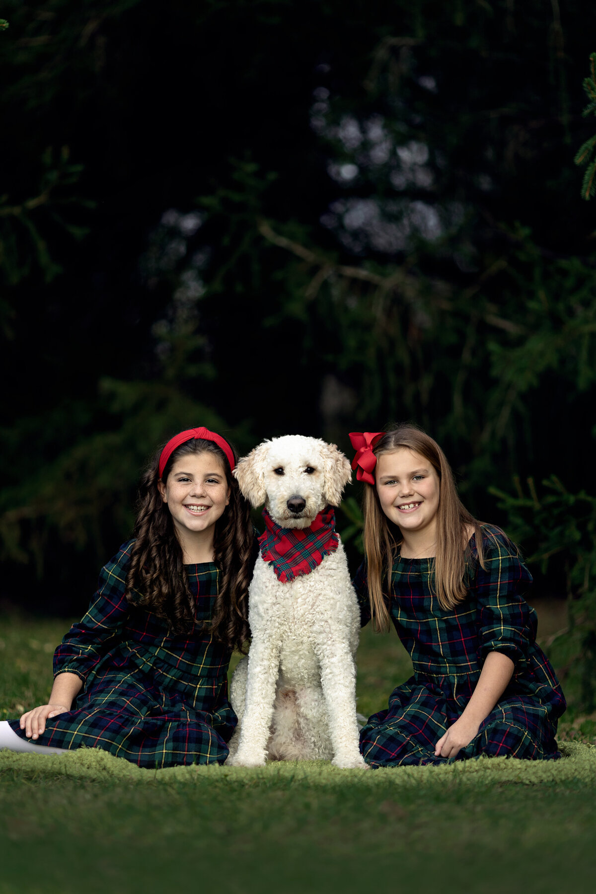 Two young sisters sit in matching dresses in a lawn with a white labradoodle wearing a scarf