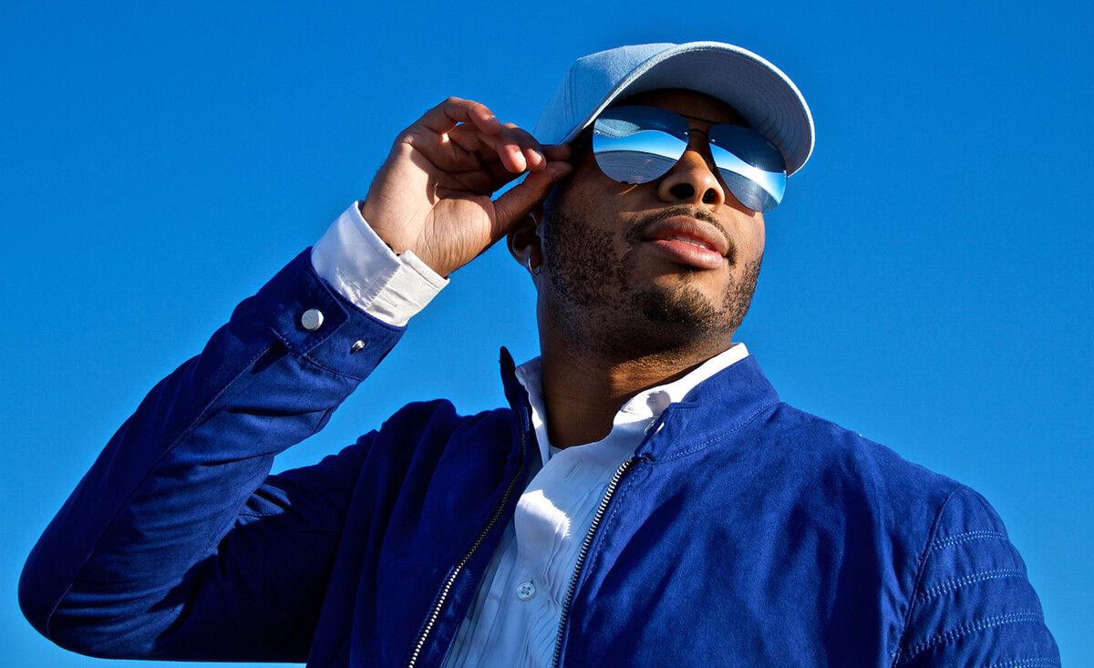 Male musician portrait Lance Ashley wearing blue jacket white baseball hat hand up holding sunglass arm with blue sky background