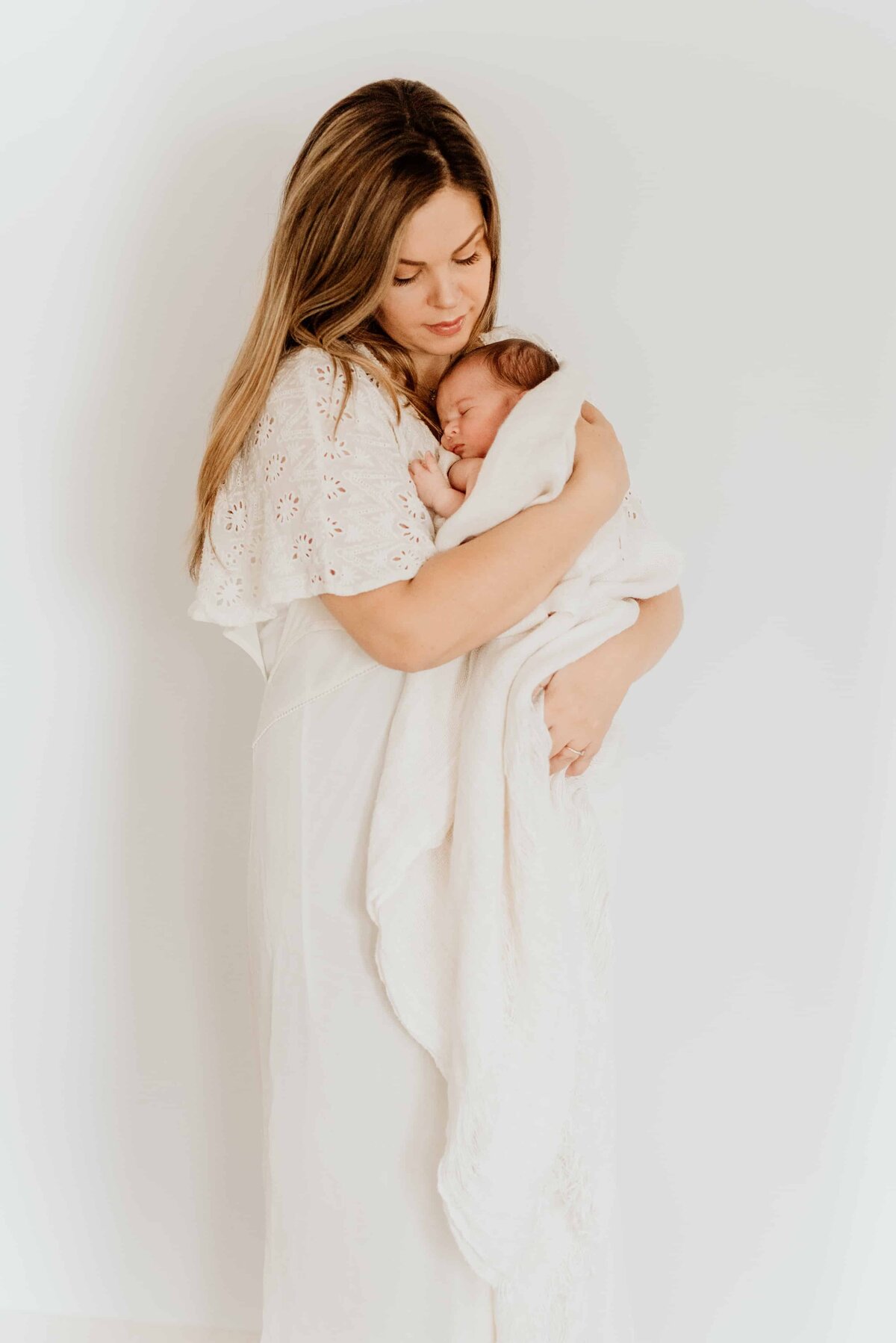 En mor står foran en hvit vegg og holder sin nyfødt datter i et teppe i armen under nyfødtfotografering hjemme.
