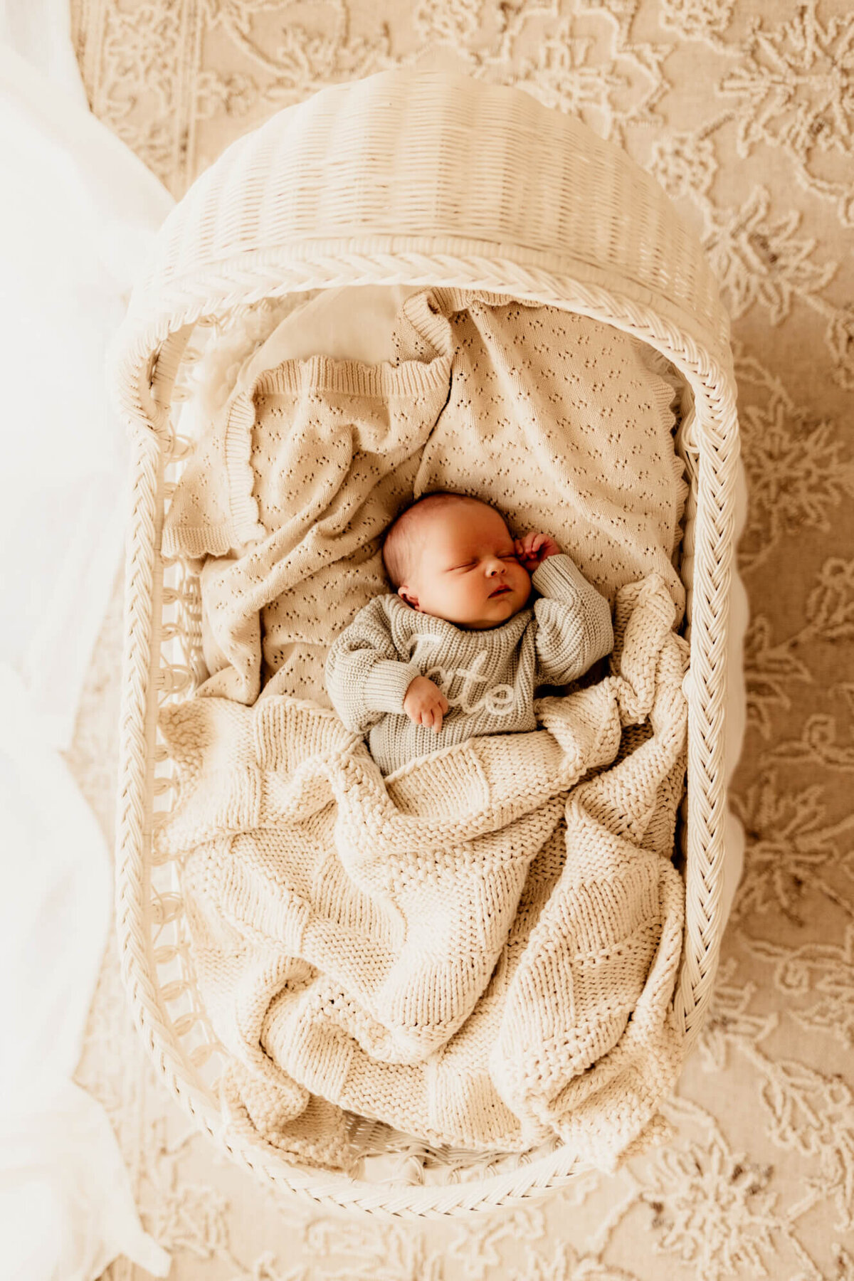 Baby boy wearing a sage blue sweater in a white wicker bassinet sleeping for a lifestyle newborn photo session in Oklahoma City.