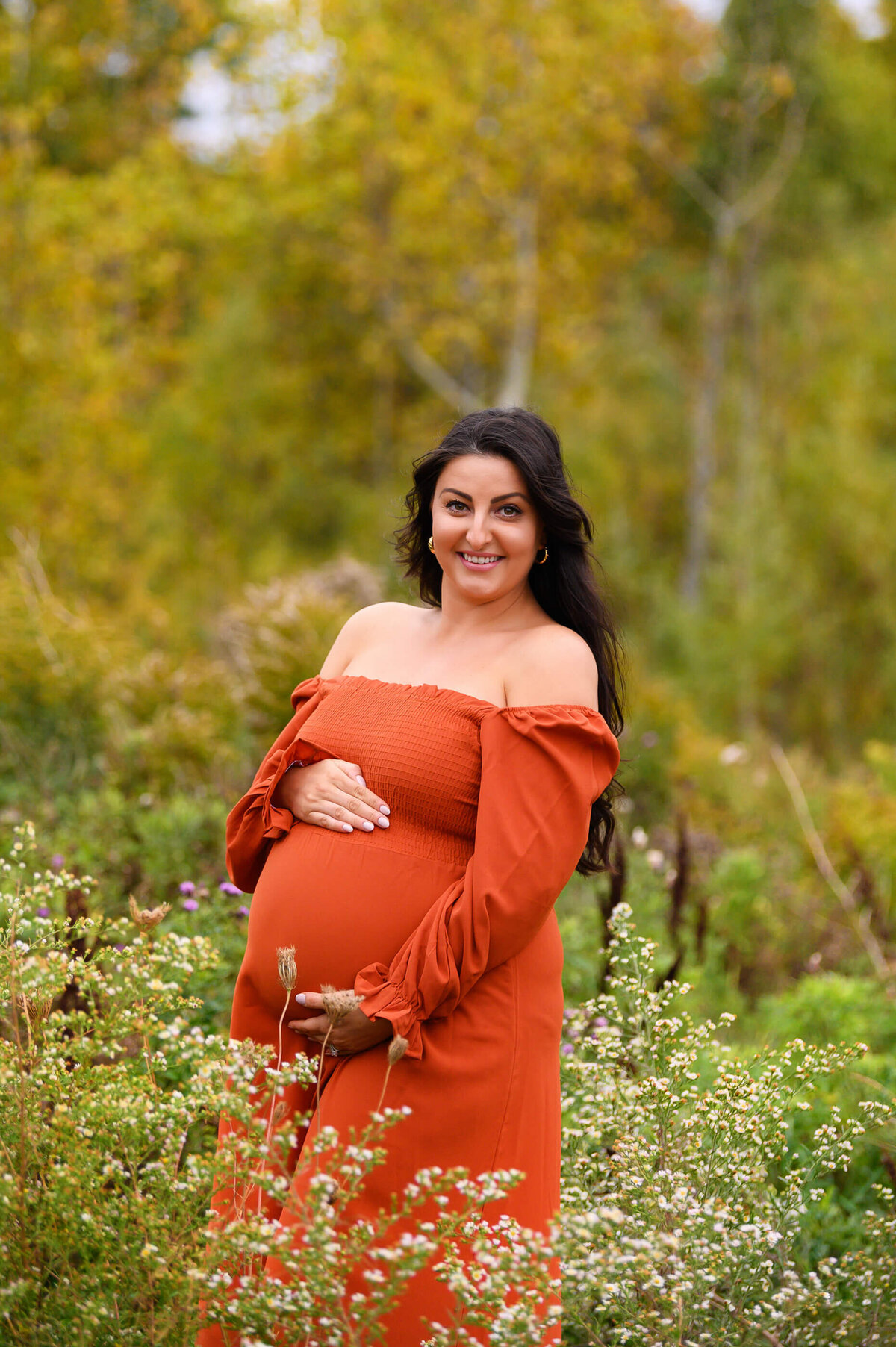 Gorgeous pregnant woman smiling cradling her belly wearing an orange maternity dress for her Toronto Fall Maternity Session