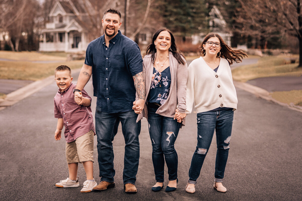 A family with older children holds hands and walks down a street in their neighborhood during an at home family photo session by Minneapolis photographer, Kate Simpson Photography.