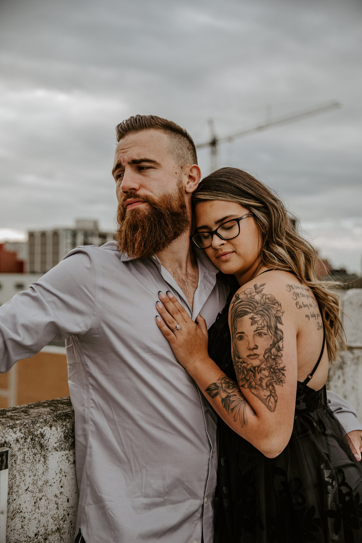 Downtown London, ON rooftop parking garage engagement photos. Man is casually leaning against the rooftop half-wall looking off in the distance. His fiance is leaning into him, resting her head on his shoulder and hand on his chest. Her arms have black outline tattoos.