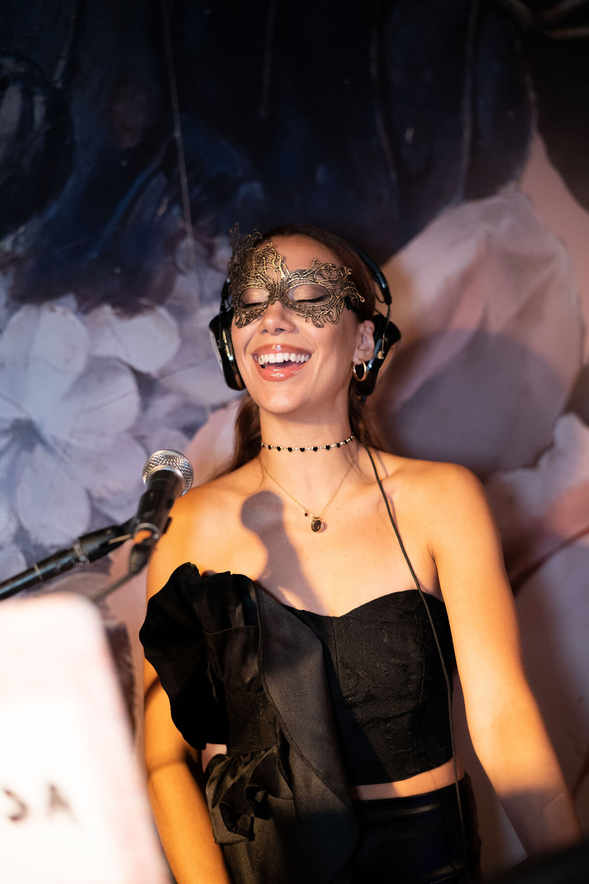 The photo was taken at a tented wedding in Virginia by the talented photographer Elisabetta Marzetti, with videography by Clayton Films. In the image, a woman with long flowing hair is captured smiling as she dons an ornate black mask over her eyes. Her outfit consists of a stylish black off-the-shoulder top adorned with delicate ruffles, paired with a fashionable choker necklace. The woman stands confidently in front of a microphone, her head adorned with headphones, set against a beautiful floral backdrop, creating a captivating and elegant scene.