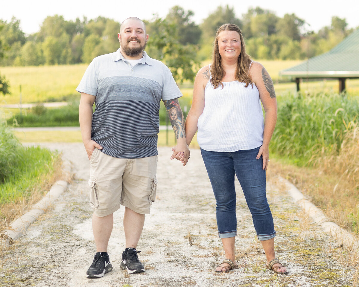 Husband and wife standing on a rocked path next to each other holding hands at a wildlife sanctuary.  Dad has a shaved head and dark brown beard, wearing a striped blue shirt, khaki shorts, black shoes, and smiling at the camera.  Wife has long brown hair, wearing a sleeveless white shirt, blue jeans, and brown sandals, smiling at the camera