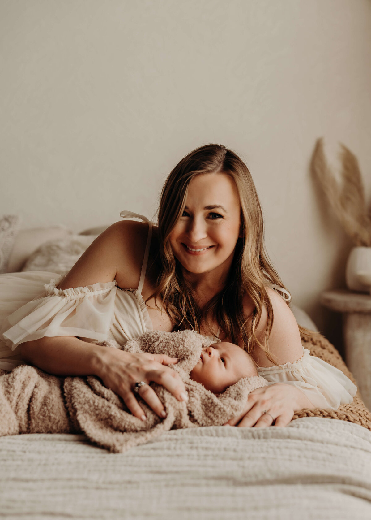 mom smiles at the camera while she lays on a bed holding her newborn
