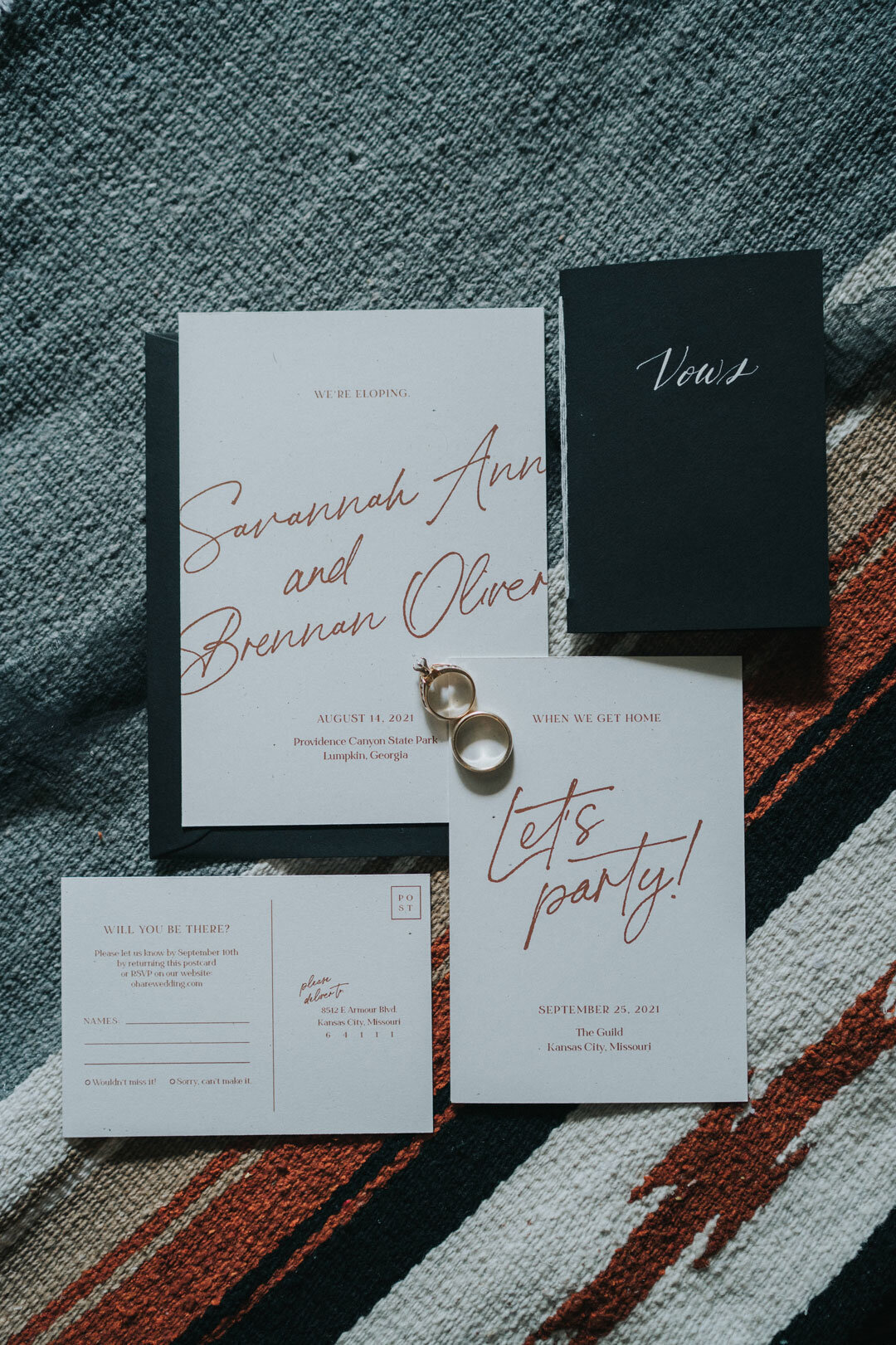 White wedding invitation with red script next to rings and black vow book