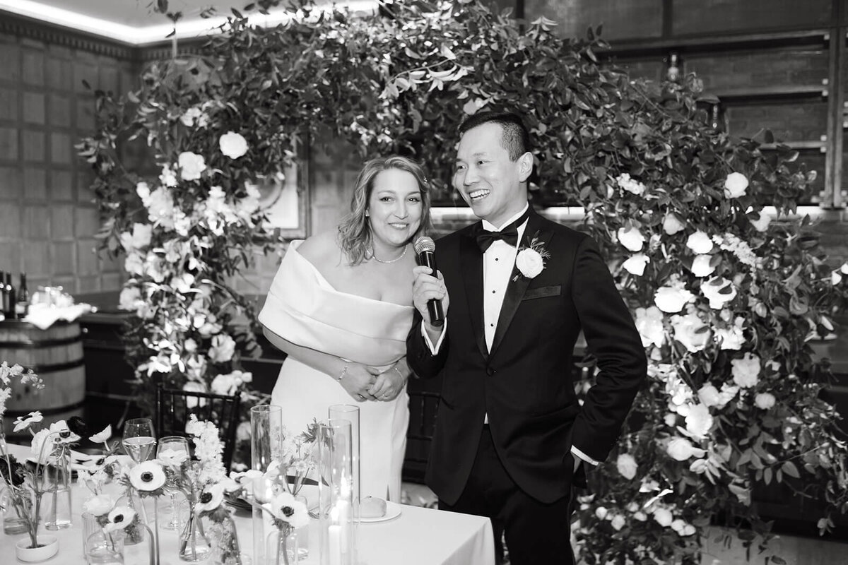 Bride and Groom stand and speak into a microphone to thank their guests among a flower filled room at the St. Regis.