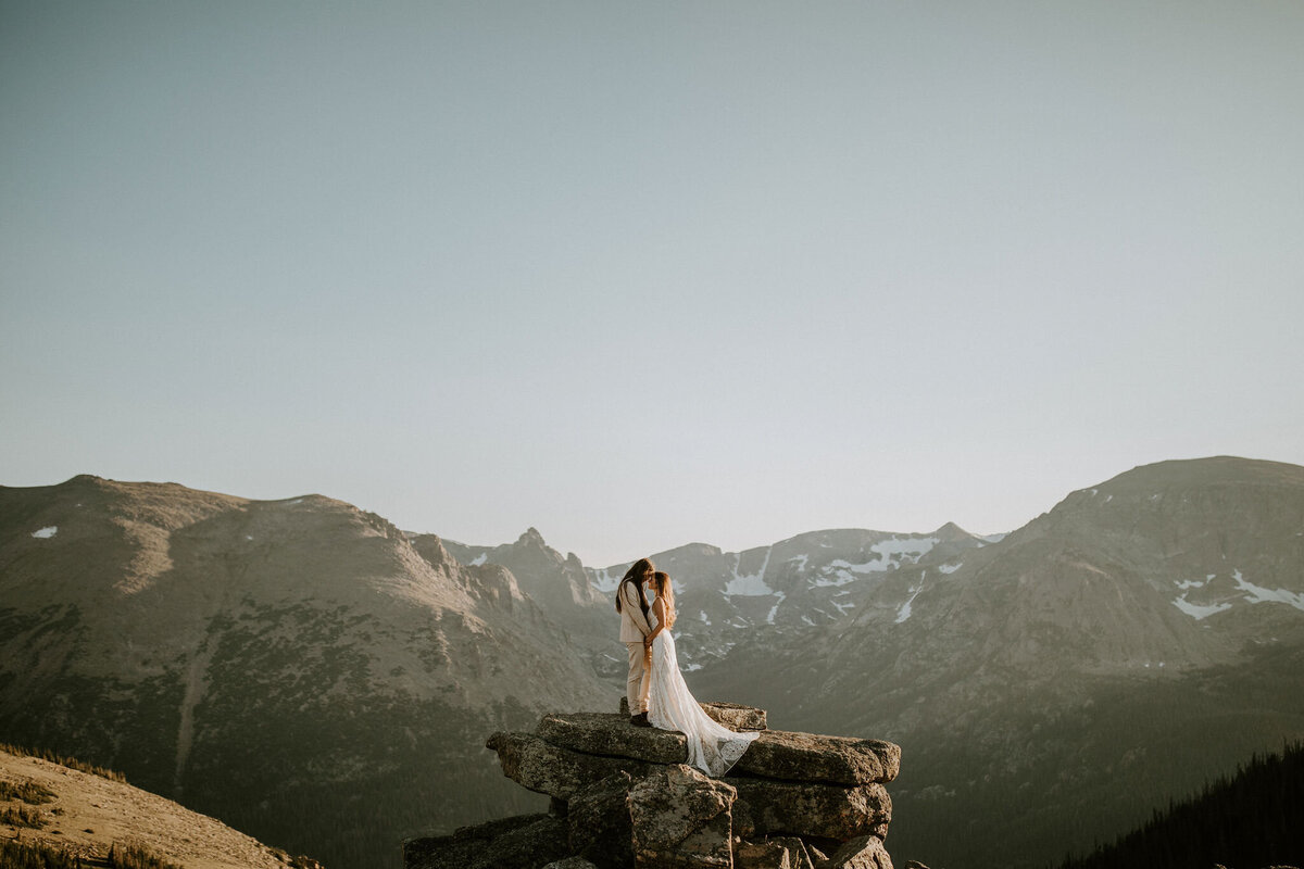 bride and groom wearing a white wedding gown and ivory tuxedo pose holding hands against mountain view