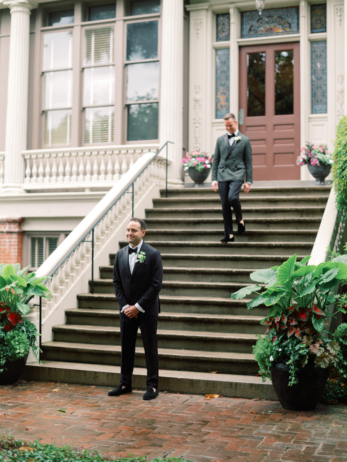 This image captures two elegantly dressed grooms in a picturesque wedding first look, showcasing love and celebration amidst beautiful greenery and architecture.
