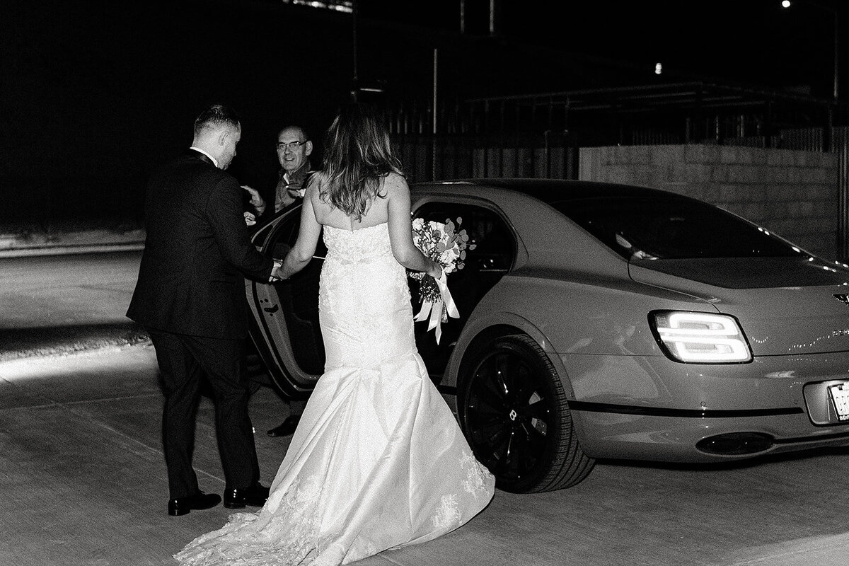 The bride, holding her bouquet, is escorted to a luxury car at night as she prepares to depart, with the groom holding the door open in a classic wedding send-off scene.