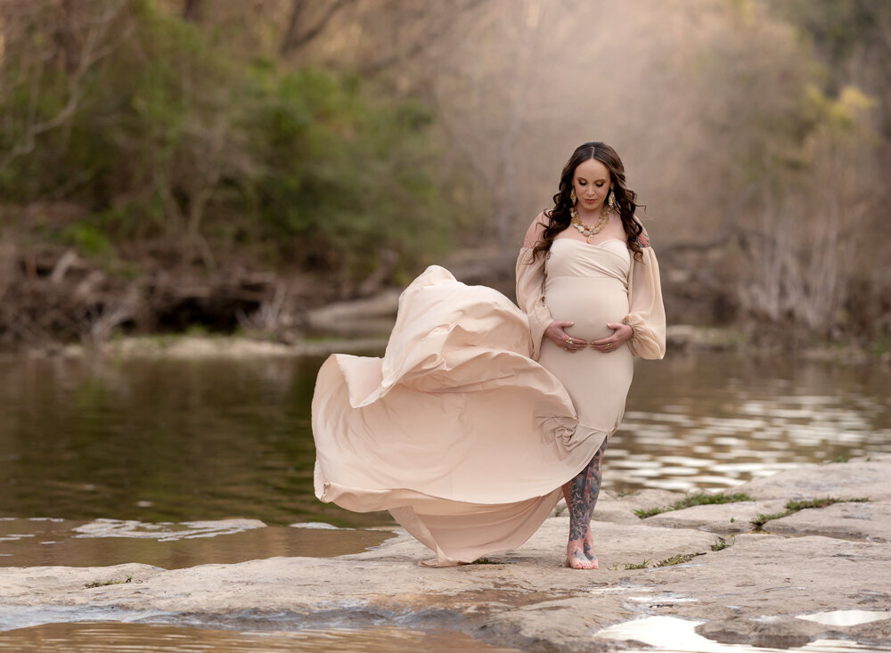 Maternity photo in Leander, TX of a barefoot mom-to-be standing by a calm stream, her soft beige gown flowing in the breeze.
