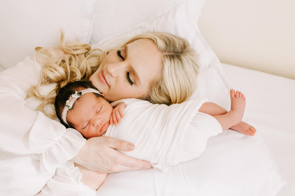 sweet portrait of mom with blonde hair holding newborn daughter. they are laying on the white bed in portland portrait studio