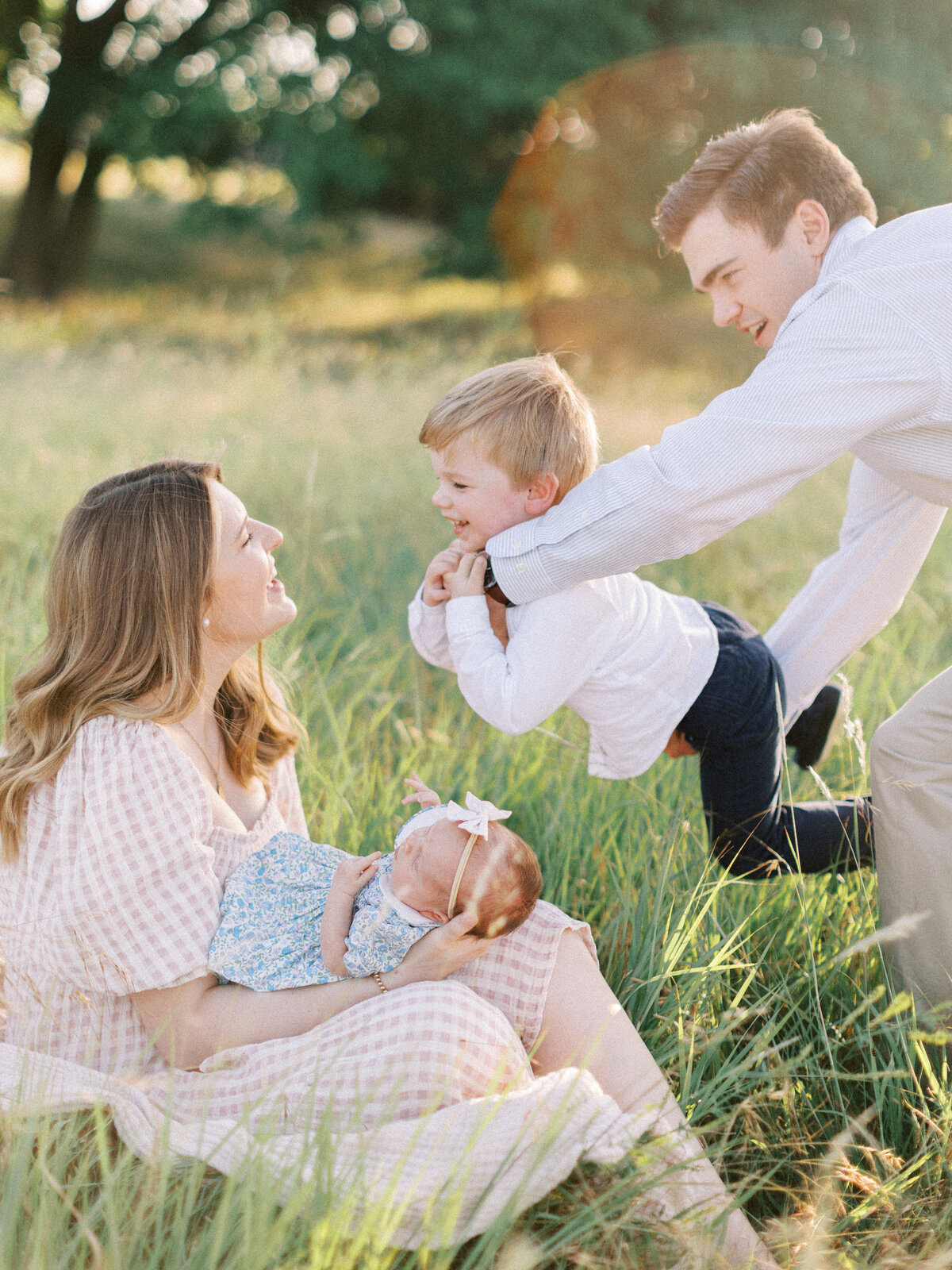 family of four plays in tall grass with mom holding newborn and dad holding toddler son