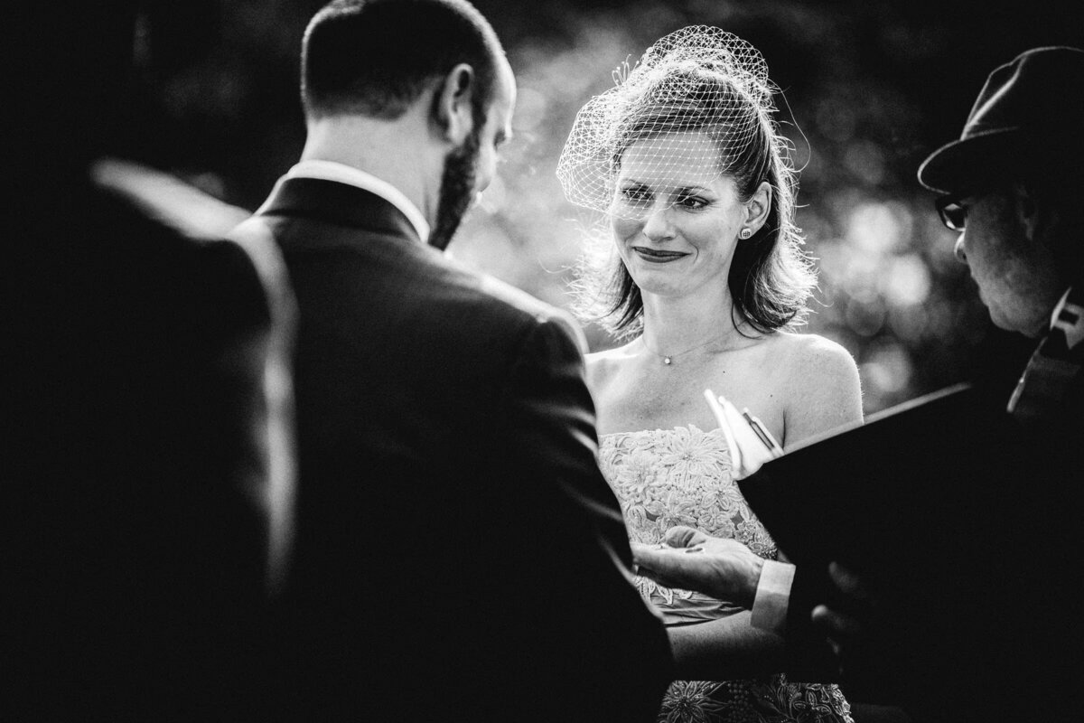 A candid black and white photo captures a bride's emotional expression as she exchanges vows with the groom, an officiant beside them.
