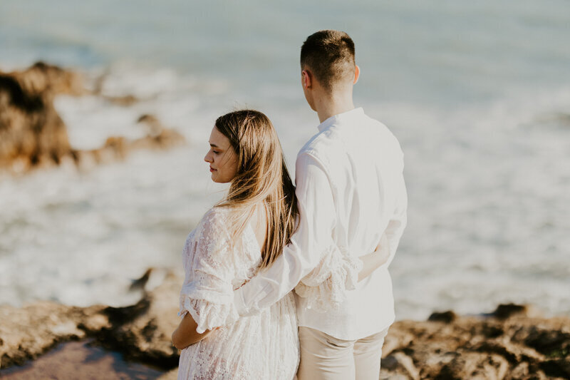 Homme, bras autour de la taille de sa femme, tous deux habillés en blanc, observant un décor de mer et de rochers, posant pour Laura Termeau photographie.