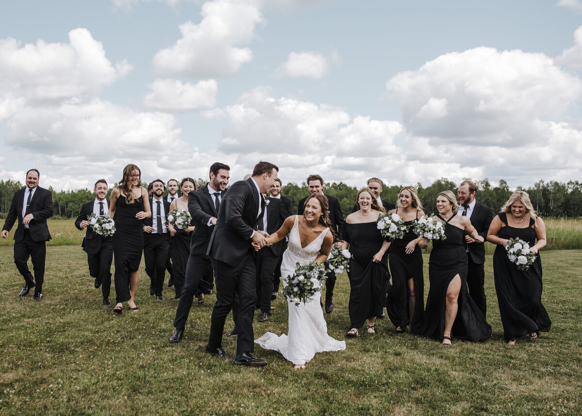 A joyful bridal party walking together on a grassy field, with the beaming bride and groom holding hands at the forefront, celebrating their special day taken by jen Jarmuzek photography a Minneapolis wedding photographer