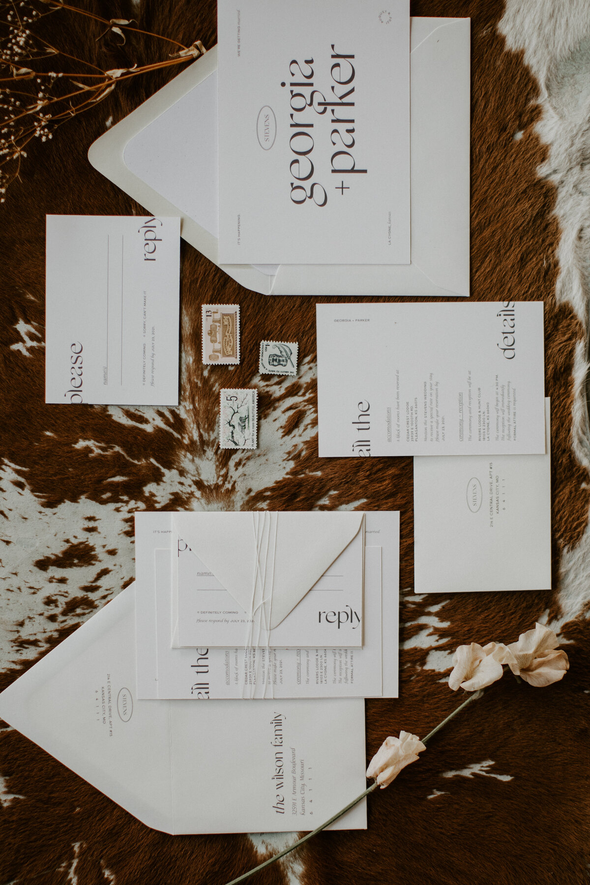 Various white wedding stationery with brown font and a dried stem and flower atop brown and white cow hide.