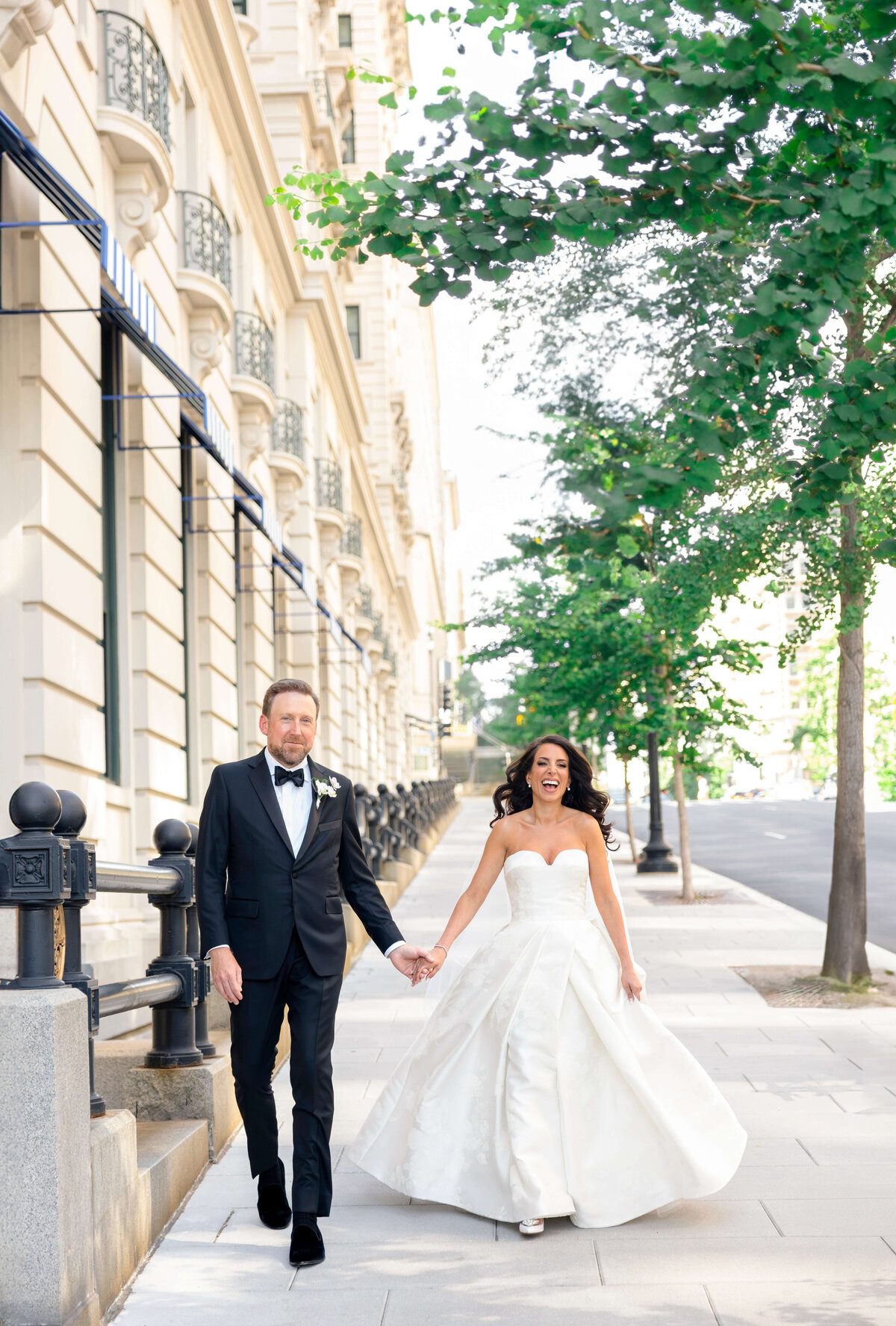 A bride and groom hold hands and walk down a city sidewalk. The groom wears a black tuxedo while the bride wears a strapless white gown. Trees line the street, and an ornate building is in the background.