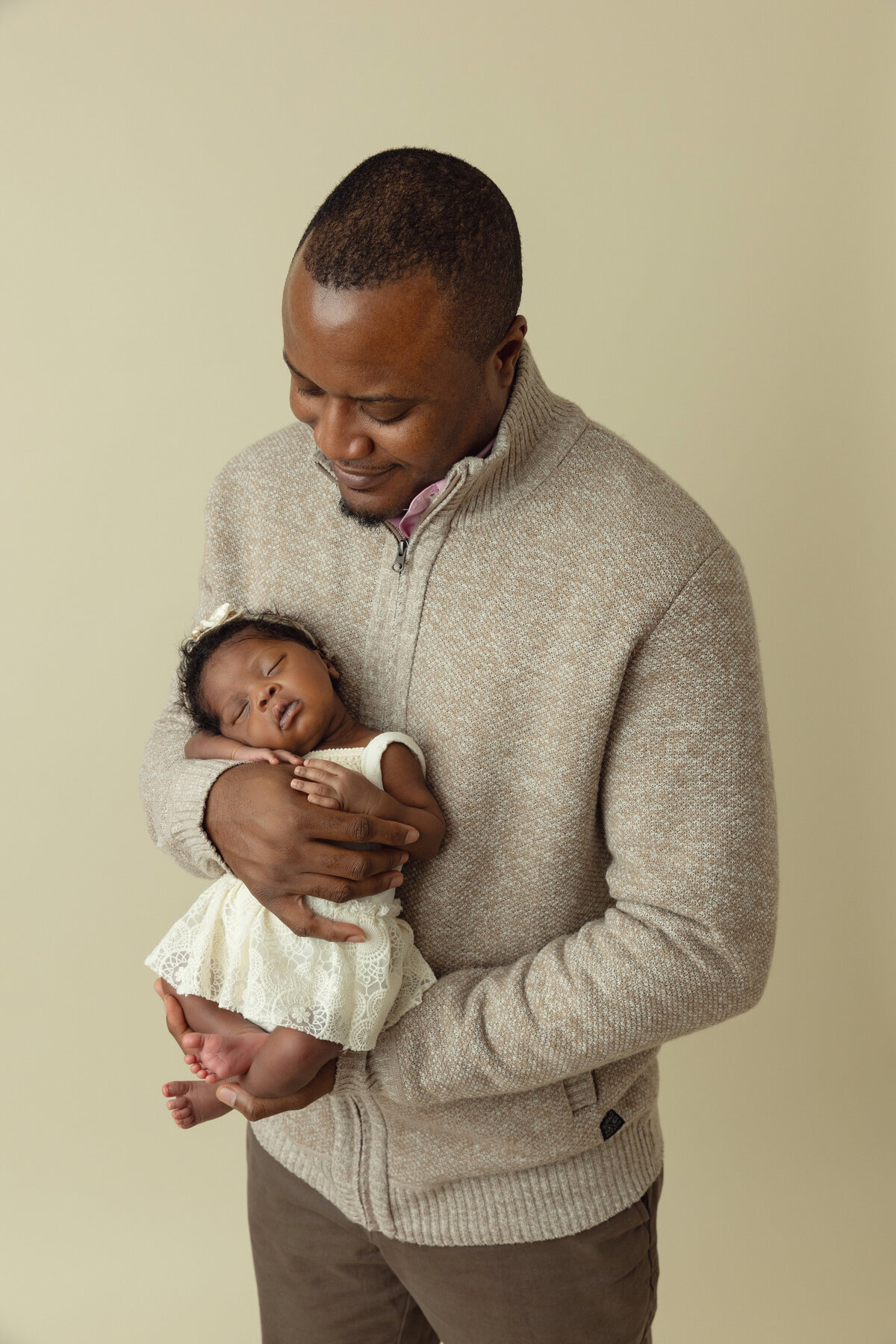 A happy dad in a grey sweater holds his sleeping newborn daughter in a white dress